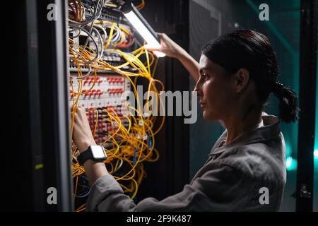 Side view portrait of female network engineer connecting cables in server cabinet while working with supercomputer in data center Stock Photo