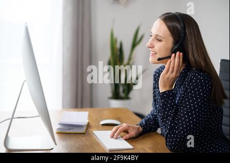 Friendly and cheerful woman wearing headset takes a call, using trendy PC for video connection with customer or colleagues on the distance, a female with hands free device at the workplace Stock Photo