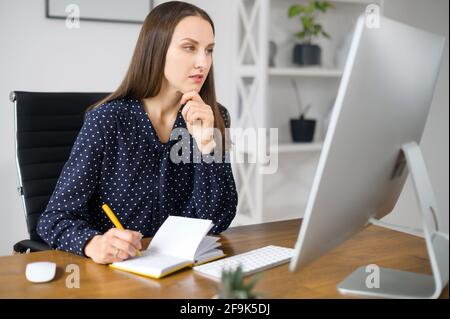 Thoughtful and concentrated female employee takes notes in notepad sitting at the desk and watching online webinars, business woman involved online conference writes down main ideas Stock Photo