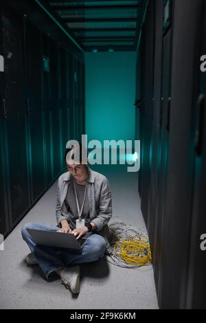 Vertical portrait of female data engineer using laptop while sitting on floor in server room and setting up supercomputer network, copy space Stock Photo