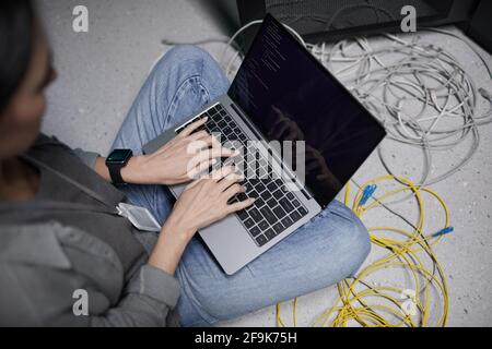 High angle closeup of female data engineer using laptop while sitting on floor in server room and setting up supercomputer network, copy space Stock Photo