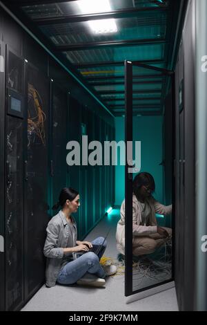 Vertical portrait of two female data engineers using laptop in server room and setting up supercomputer network, copy space Stock Photo