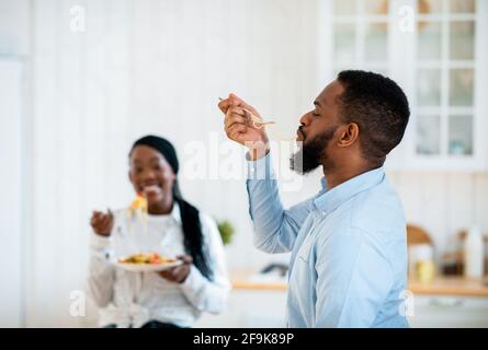 Delicious Meal. Hungry African American Man Slurping Long Spaghetti In Kitchen Stock Photo