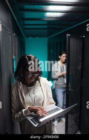 Vertical portrait of female data engineer using laptop in server room while setting up supercomputer network, copy space Stock Photo