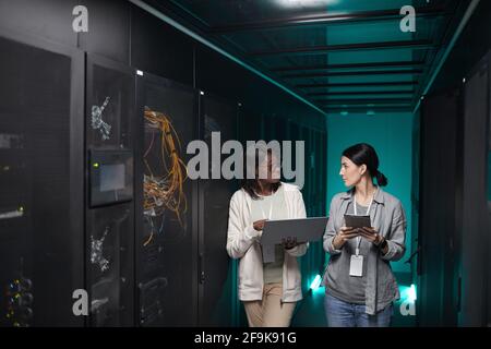Portrait of two young women using laptop in server room while setting up supercomputer network, copy space Stock Photo