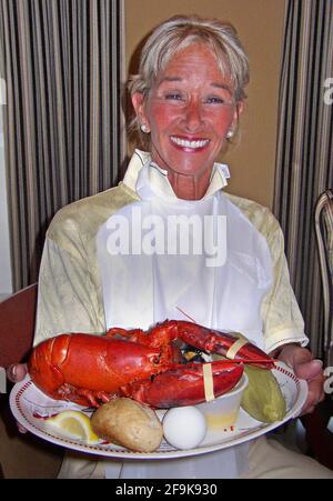 A female diner happily shows off an American lobster (Homarus americanus) that dominates the paper plate that holds a popular meal known as a “lobster boil.” The food features the large crustacean and side dishes that often include corn-on-the-cob (here in its husk), red-skinned potatoes, hard-boiled eggs, dinner rolls and clams, plus melted butter in a small container and slices of lemon. Found along the northern coast of North America, this delicious seafood is known variously as Maine, Canadian, Atlantic, Northern and true lobster. Normally greenish-brown in color, they turn red when boiled Stock Photo