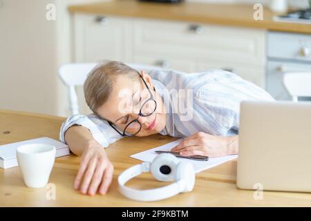 Middle aged tired woman wearing glasses sleeping on wooden table in kitchen while working or studying on laptop online from home. Senior people and technologies, remote work concept Stock Photo
