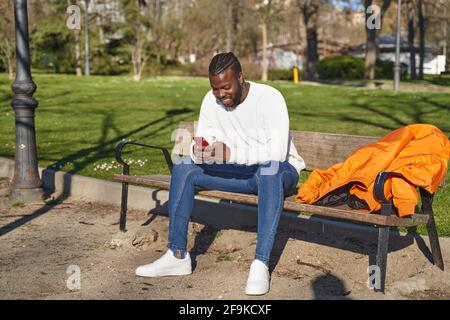 Young African American man sitting on a park bench sending a message. Concept of new normal. High quality photo Stock Photo