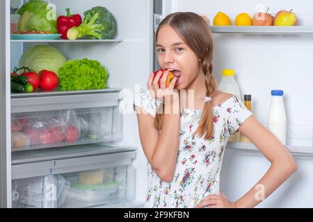 Beautiful young happy teen girl holding fresh red apple while standing near open fridge in kitchen at home Stock Photo