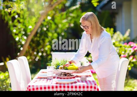 Woman setting table outdoors. Garden summer fun. BBQ in sunny backyard. Senior lady at lunch. Party decoration. Female with fruit outdoor. Stock Photo