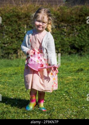 Toddler in a dress with wellington boots watering daisies, UK Stock Photo