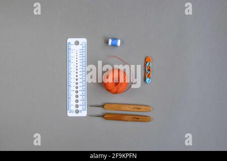 Set for needlework, knitting - two crochet hooks, round ball of orange thread, measuring ruler, pins and counter on gray background. Top view. Stock Photo