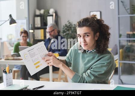 Portrait of young man looking at camera while sitting at the table and pointing at business plan at office Stock Photo