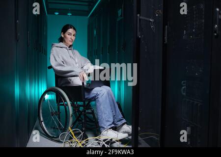 Full length portrait of disabled woman looking at camera while using laptop in server room, accessible job concept, copy space Stock Photo