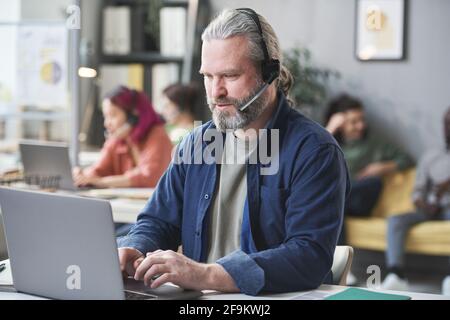Mature bearded man in headphones working on laptop at the table in call center Stock Photo