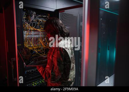 Back view at African-American woman managing computer network while working with supercomputer in server room lit by red light, copy space Stock Photo