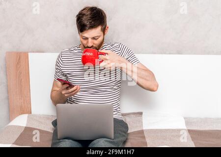 Young man working on a laptop and drinking coffee from a red big cup on the bed at home. Working remotely Stock Photo