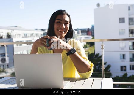 Smiling mixed transgender woman man sitting at table on sunny terrace using laptop holding coffee Stock Photo
