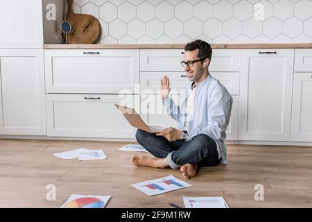 Attractive male freelancer using laptop, waving to colleagues on video meeting, smiling. Successful stylish businessman having an online conference Stock Photo