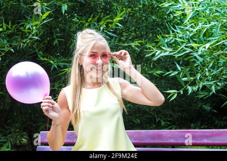 young woman sits on a bench in pink sunglasses and holds a pink balloon in her hand. The concept of happiness and positive emotions. Joy. Stock Photo
