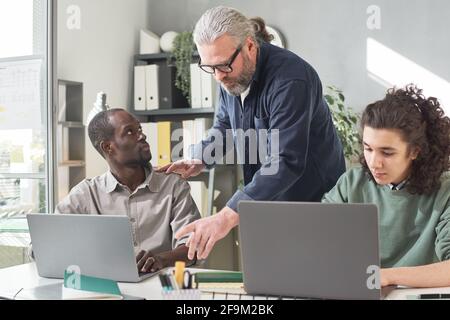 Young people sitting at the table and studying on computers with teacher talking to them Stock Photo