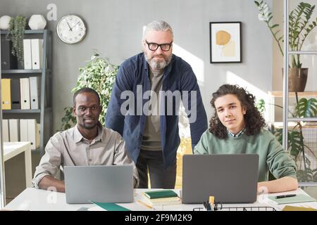 Portrait of mature teacher looking at camera with his two students sitting at the table with computers Stock Photo