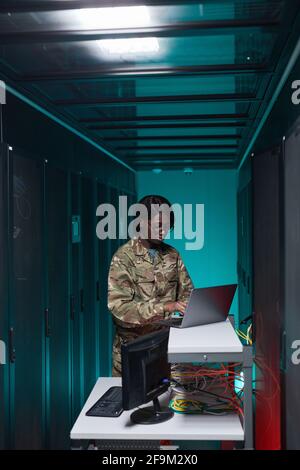 Vertical portrait of young African-American woman wearing military uniform using computer while setting up network in server room Stock Photo