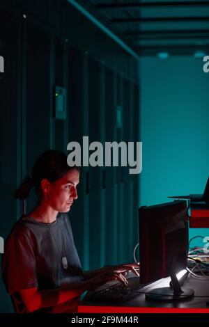 Vertical side view portrait of female IT engineer using computer while working in server room lit by blue light, copy space Stock Photo