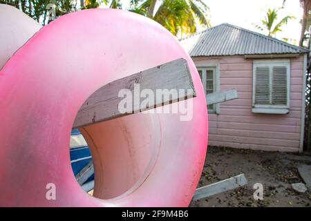 Pastel pink inflatable pool ring doughnut hanging on a wooden rack containing paddle boards and kayaks in front of a pink wooden boat house. Stock Photo