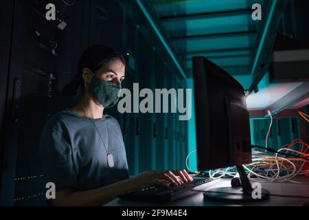 Portrait of female IT engineer wearing mask while using computer and working in server room lit by blue light, copy space Stock Photo