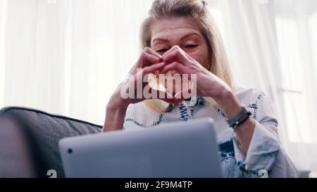 Elderly woman using laptop for a video call. Sending heart to her family. High quality photo Stock Photo