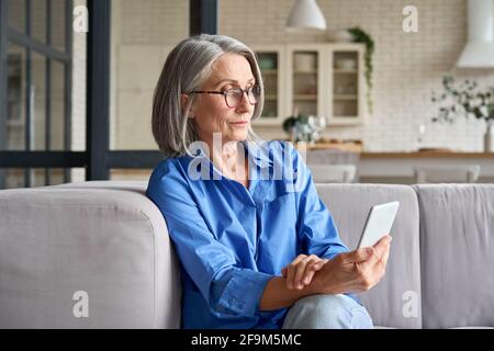 Calm mid age 60 woman at home using cellphone reading watching online video. Stock Photo