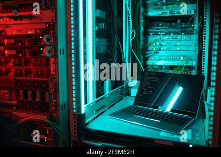 Background image of opened laptop by server cabinet in data center lit by blue light, copy space Stock Photo