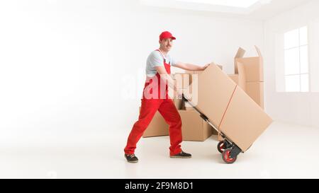 full length of young caucasian deliveryman with hand truck, transporting cardboard boxes. moving house Stock Photo