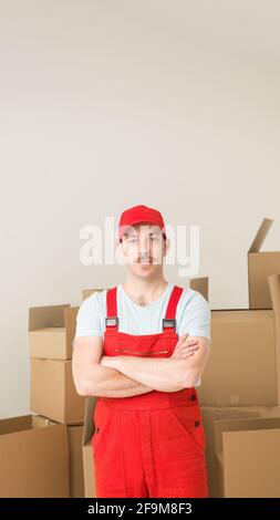 young confident delivery man standing, holding hands crossed against his chest, with cardboard boxes in background Stock Photo