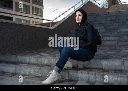 Confident woman photographer holding camera in her hands sitting on steps outdoors. Beautiful mid aged woman in hood looking into the distance Stock Photo