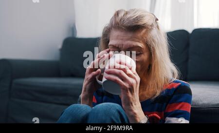 Lonely old woman drinking hot beverage while sitting on the floor in front of the couch. High quality photo Stock Photo