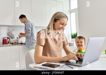 Happy mom working at home using laptop while funny kid son playing. Stock Photo