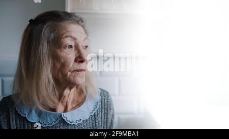 Sad lonely old gray haired woman looking through the window. Vulnerable person in quarantine. High quality photo Stock Photo