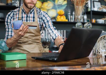Close up of caucasian seller wearing checkered shirt and beige apron typing on laptop while holding discount card. Concept of loyalty for regular customer at store. Stock Photo