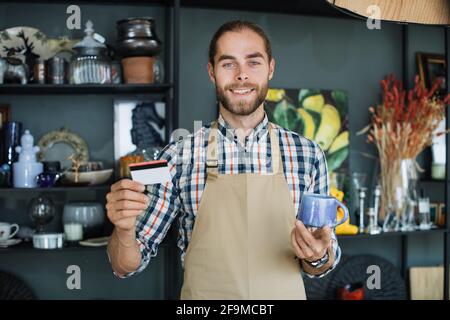 Positive bearded man holding ceramic cup and discount card in hands while posing at decor shop. Caucasian salesman in beige apron smiling and looking at camera. Stock Photo