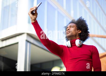Young African American man taking a picture of himself with a smart phone. He is outdoors next to a modern building. Space for text. Stock Photo