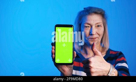 Old woman holding smartphone with green screen and showing thumb up. High quality photo Stock Photo