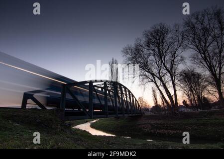 Old rusty truss bridge with moving freight train over the Orljava river. Moving train late in the evening. Rays of light. Evening arrival of the train Stock Photo