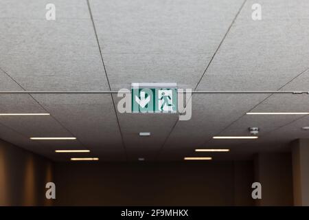 green emergency exit sign on a white ceiling with neon light on, with fire alarm in blurred background, by day, without people Stock Photo