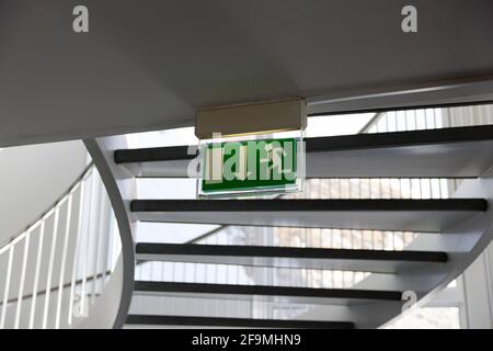 green emergency exit sign under a spiral staircase with railing, by day, without people Stock Photo