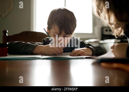 Young boy doing homework with a pencil at kitchen table with sister Stock Photo