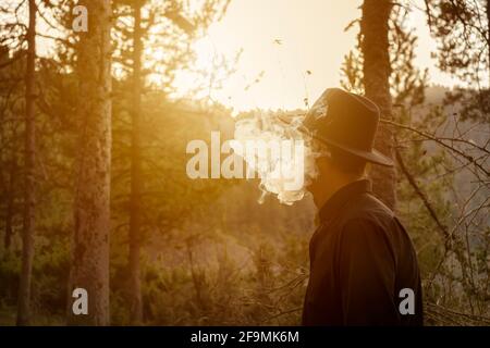 Cowboy smoking in the forest.Male in black hat and dark shirt with cloud of smoke at sunset.Beautiful creative woodland landscape with copy space. Stock Photo