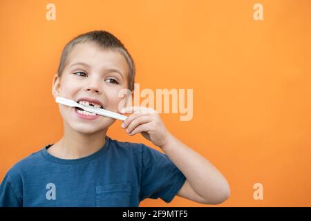 Boy without milk upper tooth in blue t-shirt holds toothbrush in his mouth on the orange background. Stock Photo