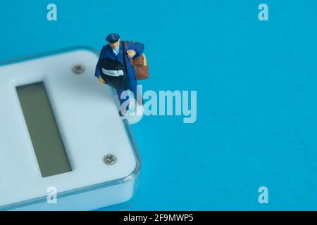 Miniature people toys conceptual photography. Calculate parcel package weight. Postman courier with scale, isolated on blue background. Stock Photo